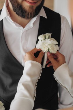 Bir adam yaka çiçeğini bir kadına yaptırıyor. Adam siyah yelek ve beyaz gömlek giyiyor.