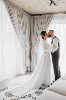 Bir gelin ve damat perdenin önünde öpüşüyor. Gelin beyaz bir elbise giyiyor ve damat siyah bir yelek giyiyor.