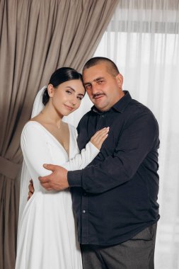 Bir erkek ve bir kadın birbirlerine sarılıyorlar. Adam siyah bir gömlek giyiyor ve kadın beyaz bir elbise giyiyor.