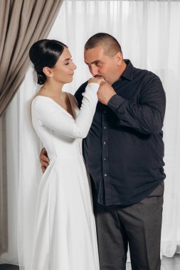 Bir erkek ve bir kadın birbirlerine sarılıyorlar. Adam kadının eline dokunuyor. Kadın beyaz bir elbise giyiyor.