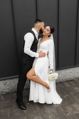 Bir gelin ve damat binanın önünde fotoğraf için poz veriyorlar. Gelin beyaz bir elbise giyiyor ve damat siyah bir takım elbise giyiyor. Çift bir buket ve küçük bir vazo tutuyor.