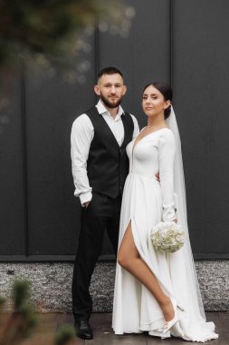 Bir gelin ve damat binanın önünde fotoğraf için poz veriyorlar. Gelin bir buket tutuyor ve damat yelek giyiyor.