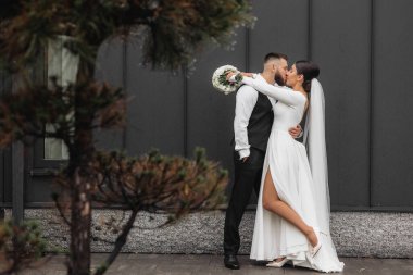Bir gelin ve damat binanın önünde öpüşüyor. Gelin beyaz bir elbise giyiyor ve damat siyah bir yelek giyiyor.