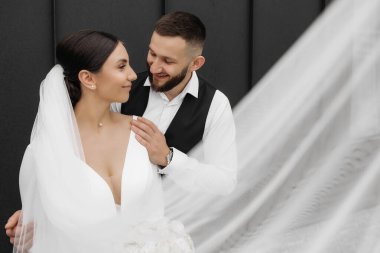 Bir gelin ve damat beyaz bir peçenin önünde fotoğraf için poz veriyorlar. Gelin beyaz bir elbise giyiyor ve damat siyah bir yelek giyiyor. Sahne mutlu ve romantik.