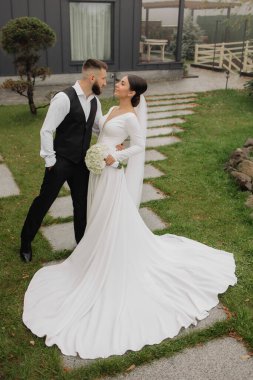 Gelin ve damat çimenli bir alanda duruyorlar. Gelin beyaz bir elbise ve damat siyah bir yelek giyiyor.