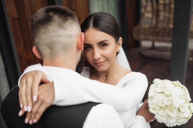 Gelin ve damat kucaklaşıyor, gelin bir buket beyaz çiçek tutuyor.