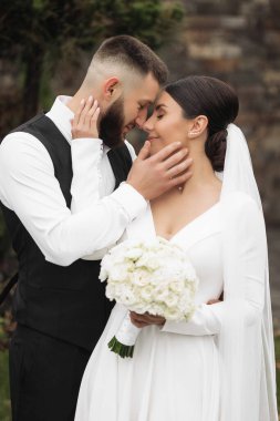 Bir gelin ve damat birbirlerine sarılıp öpüşüyorlar. Gelin elinde bir buket beyaz çiçek tutuyor.