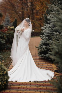 Bir gelin, bir ağacın önünde, yüzünde bir peçeyle duruyor. Elinde bir buket çiçek tutuyor.