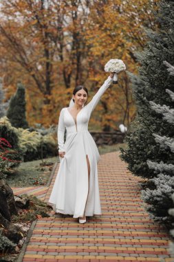 Bir gelin elinde bir buket çiçekle tuğla bir yolda yürüyor. Sahne arka planda ağaçlar olan bir parkta geçiyor. Gelin beyaz bir elbise giyiyor ve gülümsüyor.