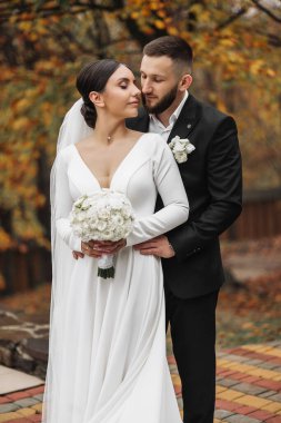 Bir gelin ve damat bir ağacın önünde fotoğraf için poz veriyorlar. Gelin bir buket tutuyor ve damat bir takım elbise giyiyor. Sahne mutlu ve romantik.