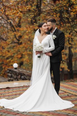 Bir gelin ve damat bir ağacın önünde fotoğraf için poz veriyorlar. Gelin beyaz bir elbise giyiyor ve damat takım elbise giyerken elinde bir buket tutuyor. Aşk ve mutluluk kavramı