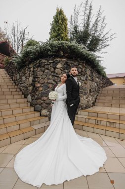 Bir gelin ve damat taş bir duvarın önünde poz veriyorlar. Gelin uzun beyaz bir elbise giyiyor ve damat takım elbise giyerken elinde bir buket tutuyor. Aşk ve bağlılık kavramı