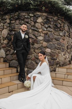 Bir gelin ve damat, taş bir duvarın önünde fotoğraf için poz veriyor. Gelin beyaz bir elbise giyiyor ve damat bir takım elbise giyiyor. Sahne mutlu ve kutlama dolu.