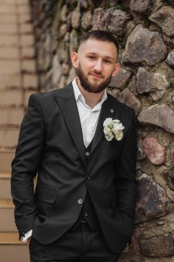 Takım elbiseli ve kravatlı bir adam taş bir duvarın önünde duruyor. O, beyaz bir gömlek ve yakasında bir çiçek giyiyor. Formalite ve zarafet kavramı