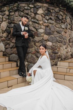 Bir gelin ve damat, taş bir duvarın önünde fotoğraf için poz veriyor. Gelin beyaz bir elbise giyiyor ve damat bir takım elbise giyiyor. Sahne mutlu ve kutlama dolu, çünkü çift evleniyor.