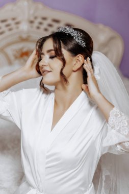 Bir kadın düğüne hazırlanıyor, duvağını takıyor ve saçını düzeltiyor. Zarafet ve entelektüellik kavramı. Kadın beyaz bir bornoz giyip taca bürünmüş.
