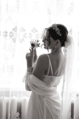 Çiçek tutan gelinlikli bir kadın. İmajın romantik ve zarif bir havası var. Kadın peçe ve kolye takıyor ve pencerenin önünde duruyor.