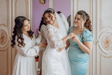 Üç kadın fotoğraf için poz veriyor, biri gelinlik giyiyor. Diğer iki kadın elbisesini düzeltmesine yardım ediyor. Sahne neşeli ve neşeli.