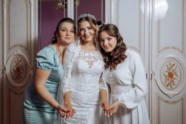 Üç kadın fotoğraf için poz veriyor, biri gelinlik giyiyor. Kadınlar özel bir durum için poz verirken sahne mutlu ve kutlama dolu.