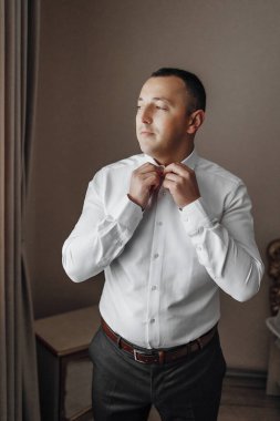 Bir adam beyaz gömlek ve gri pantolon giymeye hazırlanıyor. Kravatını düzeltiyor ve pencereden dışarı bakıyor.