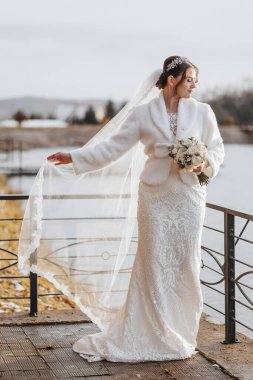 Beyaz kürk manto ve duvağıyla bir gelin köprüde duruyor. Elinde bir buket çiçek tutuyor.