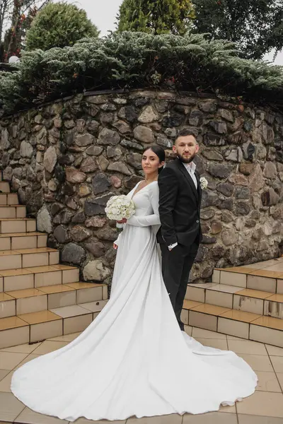 Bir gelin ve damat, taş bir duvarın önünde fotoğraf için poz veriyor. Gelin uzun beyaz bir elbise giyiyor ve damat bir takım elbise giyiyor. Aşk ve bağlılık kavramı