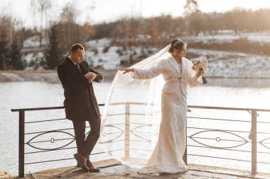 Bir gelin ve damat, bir su kütlesine tepeden bakan bir iskelede duruyor. Gelin bir buket çiçek tutuyor ve damat bir saat takıyor. Sahne sakin ve romantik.