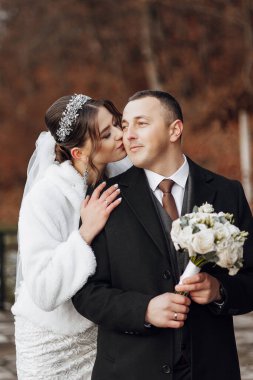 Bir gelin ve damat birbirlerinin yanaklarını öpüyor. Gelin beyaz bir ceket giyiyor ve damat siyah bir takım elbise giyiyor. Damat elinde bir buket çiçek tutuyor.