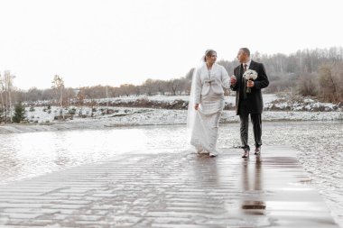 Bir gelin ve damat karda bir köprüde yürüyorlar. Gelin beyaz bir elbise giyiyor ve damat siyah bir takım elbise giyiyor. El ele tutuşuyorlar ve birlikte yürüyorlar, özel günlerinin tadını çıkarıyorlar.