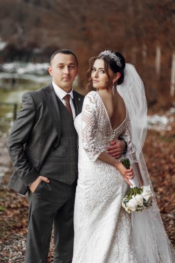 Bir gelin ve damat ormanda fotoğraf çektirmek için poz veriyorlar. Damat takım elbise giyerken gelin beyaz bir elbise ve taç giyiyor. Bir buket çiçek ve çiçek tutuyorlar.