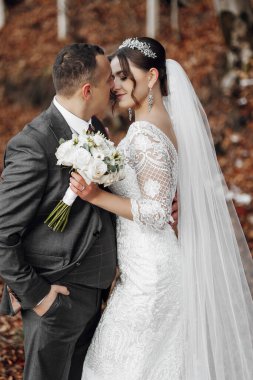 Bir gelin ve damat birbirlerine sarılıp öpüşüyorlar. Gelin beyaz bir elbise ve duvak takıyor. Yerde yapraklarla bir tarlada duruyorlar.