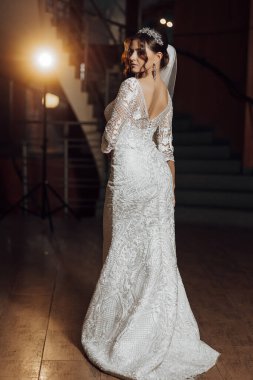 Beyaz elbiseli bir kadın bir merdivenin önünde duruyor. Bir taç ve duvak takıyor.