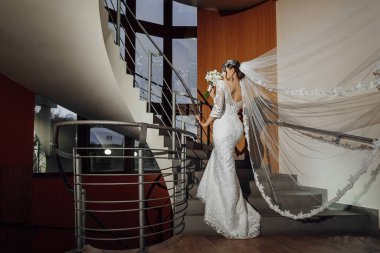 Gelinlikle merdivenlerden inen bir gelin var. Elinde bir buket çiçek ve duvak tutuyor. Gelin, düğün kıyafetlerini giydiği için, zarafet ve kutlama kavramı.