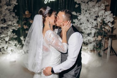 Bir gelin ve damat beyaz bir kemerin önünde öpüşüyor. Gelin peçe takıyor ve damat yelek giyiyor. Sahne romantik ve mutlu.