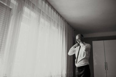 Bir adam işe hazırlanıyor, kravatını takıyor ve yakasını düzeltiyor. Oda loş bir şekilde aydınlatılmış, pencere de az miktarda ışık alıyor.