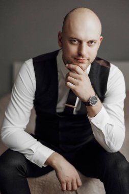 Takım elbiseli ve yelekli bir adam elleri bağlı bir kanepede oturur. Saat takıyor ve yüzünde ciddi bir ifade var.