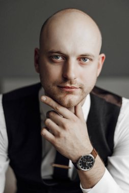 Kel kafalı ve bileğinde saat olan bir adam resim için poz veriyor. Siyah bir yelek ve beyaz bir gömlek giyiyor. Resmin profesyonel ve resmi bir havası var.