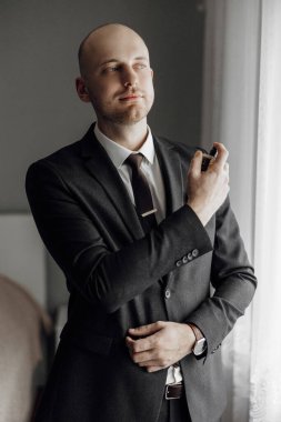 Takım elbiseli ve kravatlı bir adam parfüm sıkmaya hazırlanıyor. Resmin profesyonel ve parlak bir havası var. Pencere önünde duran adam.