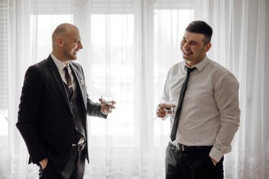 Takım elbiseli iki adam gülüyor ve bardaktan içiyor. İçlerinden biri kravat takıyor. Sahne neşeli ve dostane.