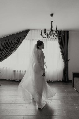 Beyaz elbiseli bir kadın üzerinde avizeyle pencerenin önünde duruyor.