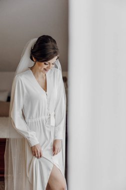 Beyaz elbiseli bir kadın pencerenin önünde duruyor. Duvak ve beyaz bir elbise giyiyor.