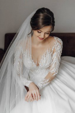 Beyaz bir gelinlik giymiş bir kadın yatakta oturuyor. Beyaz bir peçe takıyor ve elleri kucağında.