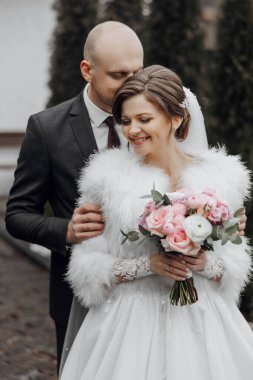 Bir gelin ve damat sarılıyor ve gelin bir buket çiçek tutuyor. Gelin beyaz bir elbise giyiyor ve damat bir takım elbise giyiyor. Sahne mutlu ve romantik.