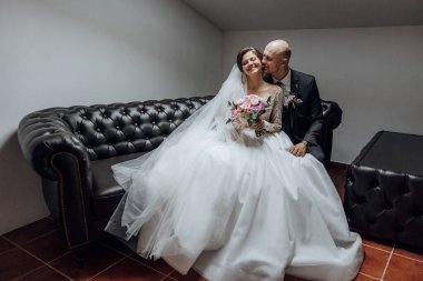 Gelin ve damat siyah bir kanepede oturuyor, gelin beyaz bir elbise ve damat siyah bir takım elbise giyiyor. Gelin bir buket çiçek tutuyor ve çift birbirini öpüyor.