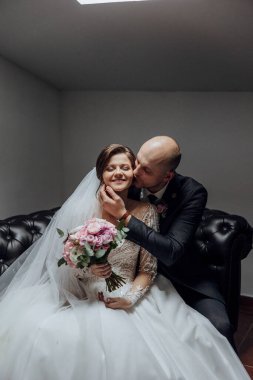 Gelin ve damat siyah bir kanepede oturuyor ve gelin elinde bir buket çiçek tutuyor. Sahne romantik ve samimi, çünkü çift birbirine sarılıp öpüşüyor.