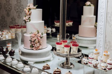 Beyaz pastalı bir masa ve üstünde pembe pasta olan gümüş bir tepsi. Masada başka tatlılar da var.