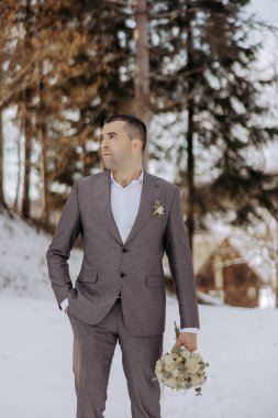 Takım elbiseli bir adam elinde bir buket çiçekle karda duruyor. Sahne sakin ve huzurlu, adam uzaklığa doğru bakıyor. Adamın resmi kıyafetlerinin birleşimi.