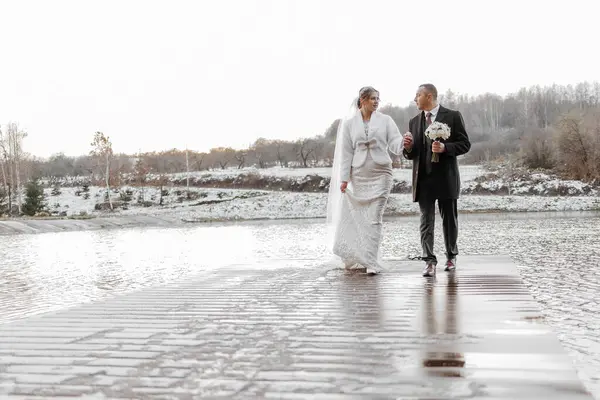 A bride and groom are walking on a bridge in the snow. The bride is wearing a white dress and the groom is wearing a black suit. They are holding hands and walking together, enjoying their special day