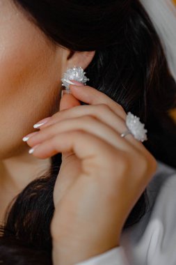 Bir kadın kulağını tutuyor ve bir çift küpe takıyor. Küpeler beyaz ve çiçek desenli.