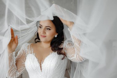 Gelinlikli bir kadın beyaz perdenin önünde duruyor. Gülümsüyor ve kameraya bakıyor.